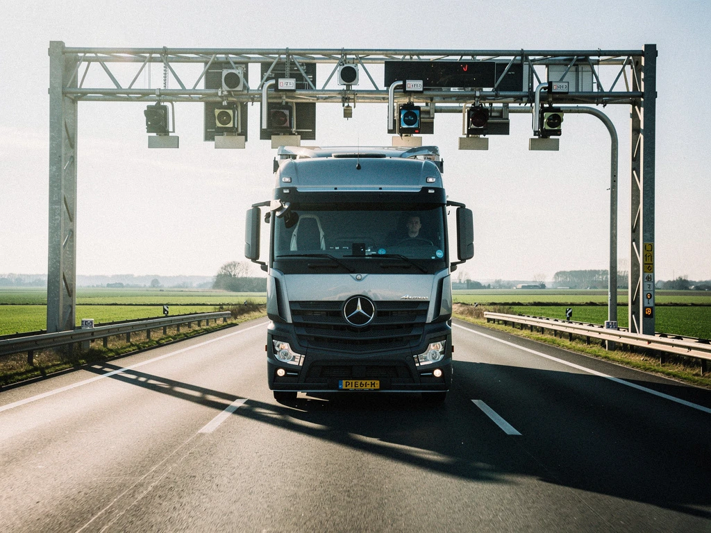 Zware vrachtwagen rijdt onder elektronische tolpoortjes op Nederlandse snelweg met groene landschap op achtergrond