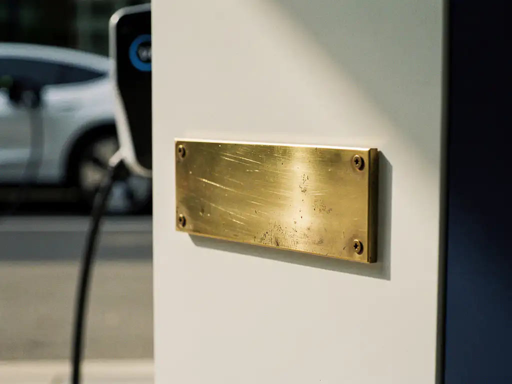 Verweerde messing naamplaat op kantoorwand vervangen door glanzend nieuw exemplaar, elektrisch voertuig op achtergrond.